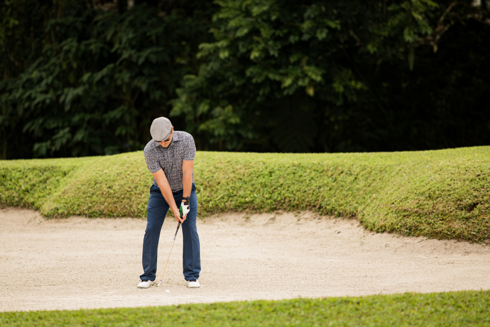 A male golfer in a flat cap and patterned shirt prepares to hit a shot from a sand bunker on a lush green course.