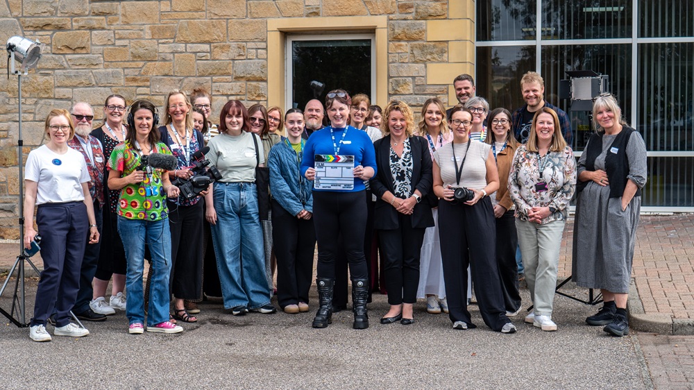 A large group of people standing in front of a building. The person in the centre holds a clapper board. A large group of people standing in front of a building. The person in the centre holds a clapper board.