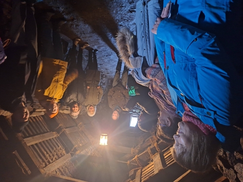 A group in warm outdoor clothing gathered inside a dimly lit, rustic building with exposed stone walls, a thatched roof, and a central hanging light. A group in warm outdoor clothing gathered inside a dimly lit, rustic building with exposed stone walls, a thatched roof, and a central hanging light.
