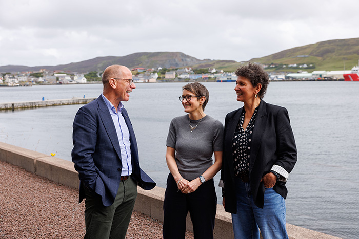 Three people in conversation outside with the sea and buildings visible behind them