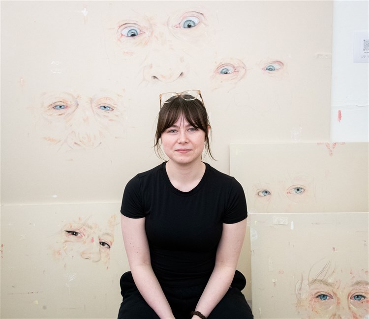 A artist sitting in their studio with art on the wall behinf them