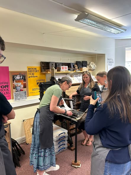 Woman wearing an apron bending over a manual printing press. Group of four people observe them working, and posters on the walls and prints on the shelves showcase different kinds of printing products