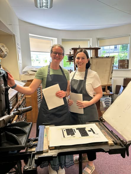 Two women wearing aprons standing next to a manual printing press
