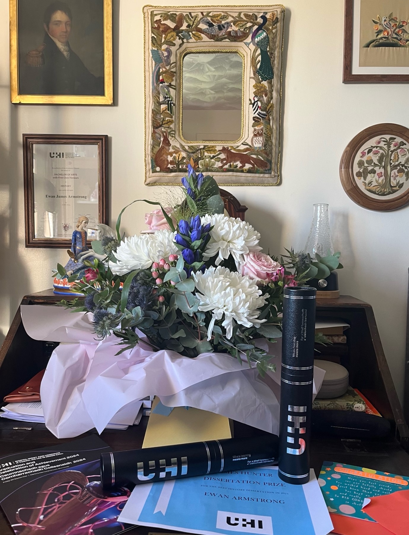 A still-life style grouping of items on a desk, with a bouquet of flowers, graduation scroll cases and UHI printouts on the table, while on the wall detailed, framed pieces of needlework, a painting and a framed graduation certificate complete the scene.