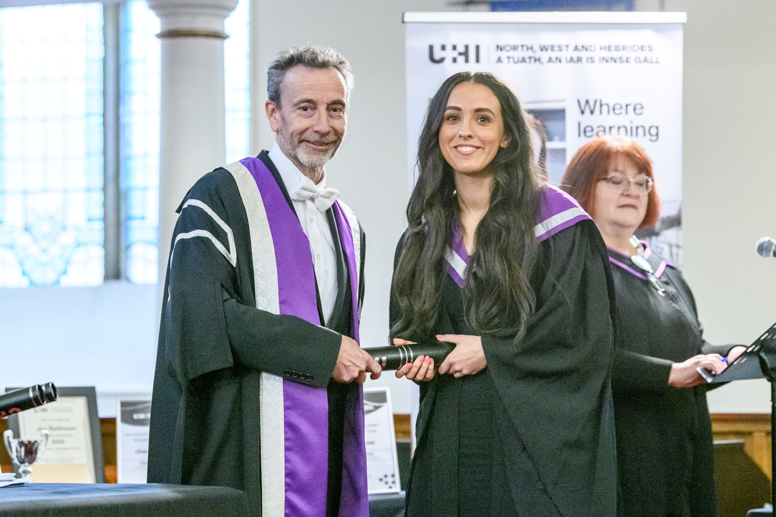 Woman and man in academic gowns and hoods both holding on to a graduation scroll and looking at the camera smiling