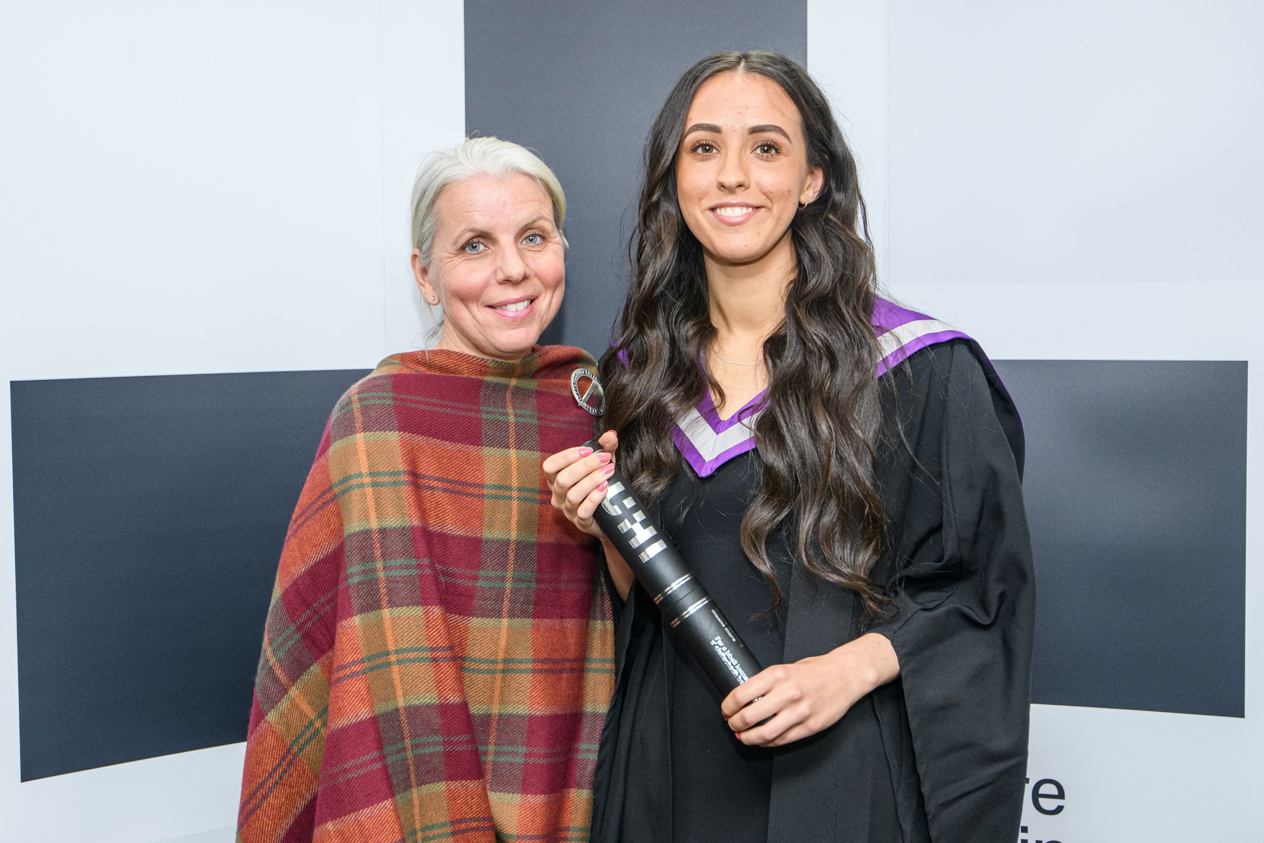 Two women smiling at the camera. The other one is wearing a graduation gown and hood, while the other wears a tartan-patterned shawl.
