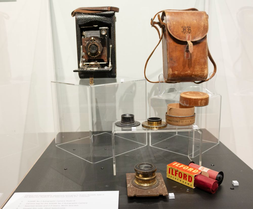 Bobbie Barwell’s No. 3 Autographic Kodak Model H, a folding bellows roll film camera, and accessories, including assorted lenses, unused rolls of film and a camera bag inscribed with the initials B.B., displayed in Bobbie Barwell: Capturing People and Places, Ashburton Art Gallery and Museum, 2022.